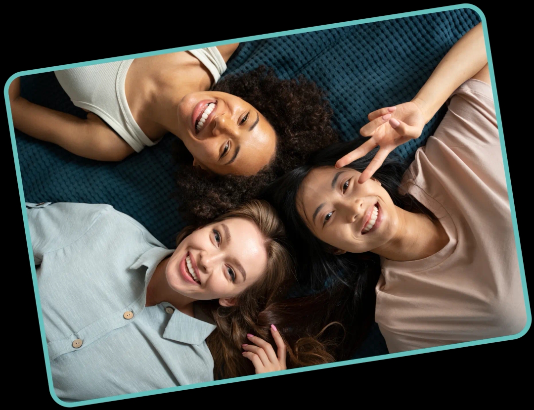 Three smiling young women lie on a dark blue surface, with one making a peace sign, framed by a tilted turquoise border.